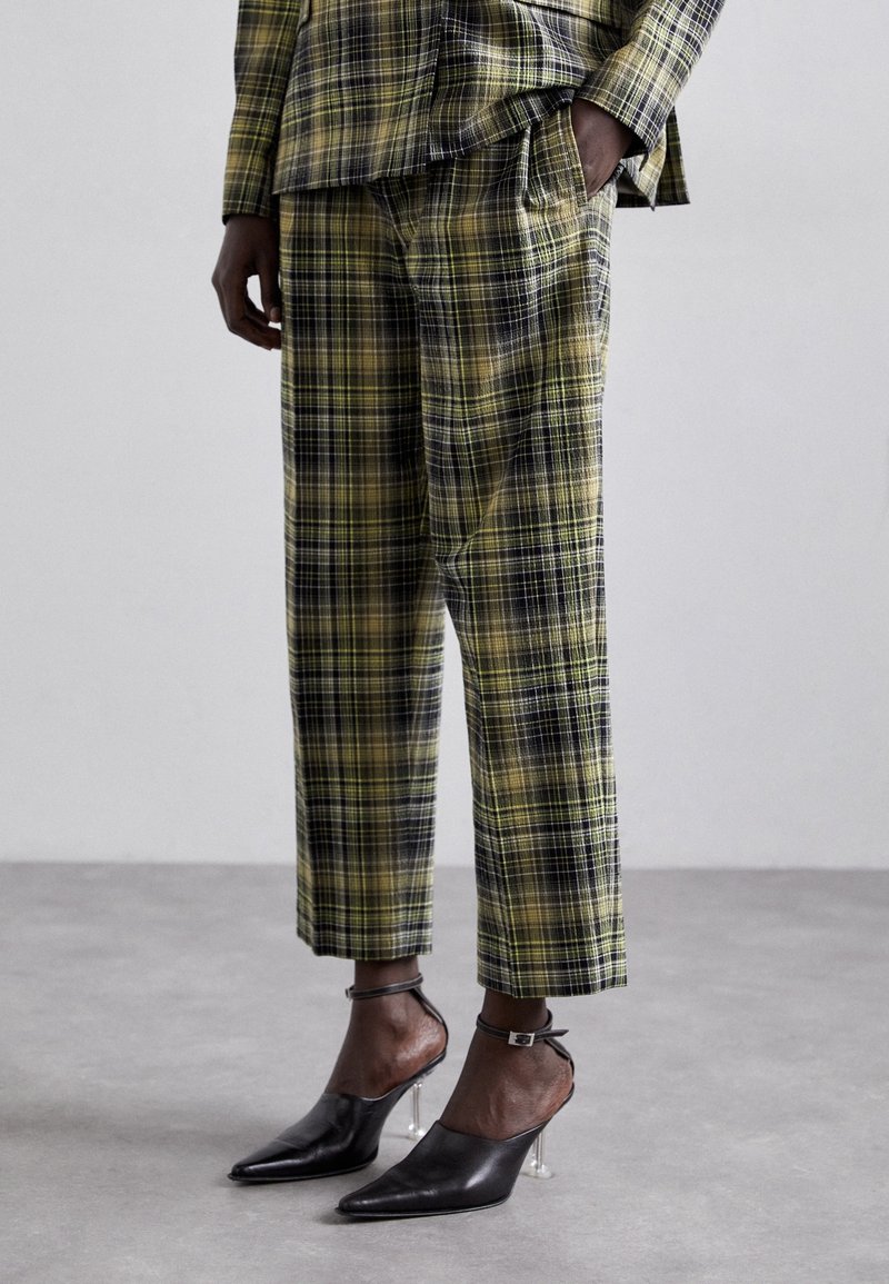 Personne portant un pantalon court à carreaux verts et noirs et un haut assorti, avec des talons pointus noirs munis de talons blocs transparents.