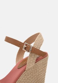 Sandale compensée avec talon en jute tressé, semelle intérieure en tissu rose, bride de cheville en tresse beige, et bride à boucle en cuir marron sur fond blanc uni.