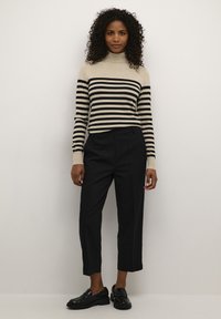 Bežs turtlenecks džemperis ar melnām svītrām, pārī ar piegrieztām melnām potīšu biksēm. Apavi ir spīdīgi melni loafers ar masīvu zoli.
