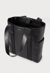 Sac fourre-tout noir en matériau lisse, doté de deux poignées plates et d'une poche latérale. "Calvin Klein" est embossé sur le devant.