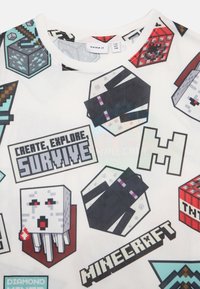 Weißes T-Shirt mit Minecraft-Themen-Grafiken, einschließlich Charakteren, Werkzeugen und Blöcken. Der Text lautet "Kreativ sein, Entdecken, Überleben."