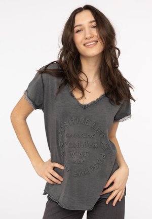 Junge Frau mit langen braunen Haaren lächelt in einem verblassten schwarzen V-Ausschnitt-Shirt mit dem Text "Positives Leben, positiver Geist" und Herzsymbolen.