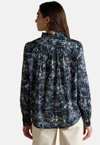 Langärmlige Bluse mit navyblauem Hintergrund und einem weißen Blumenmuster, Rüschkragen und gerafftem Rückendetail. Glatte Stofftextur.