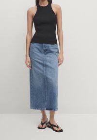 Kvinna som bär en svart ärmlös ribbad topp, ljusblå denim midi-kjol och svarta platta sandaler, stående mot en enfärgad bakgrund.