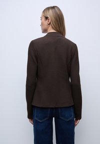 Hnědý texturovaný blazer se střihem na míru, kulatým výstřihem a dlouhými rukávy, spárovaný s tmavě modrými džíny.