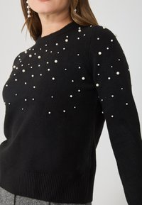 Czarny sweter z dzianiny z rozrzuconymi białymi perłowymi zdobieniami, ribbowanymi mankietami i dołem. Klasyczny okrągły dekolt i dopasowany fason.