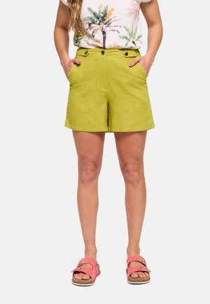 Mujer con shorts verde lima de talle alto con botones delanteros, camiseta con estampado de palmeras y sandalias rosas con doble tira, de pie con las manos en los bolsillos.