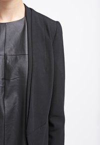 Černý přizpůsobený blazer s hladkým povrchem, s límečkem a otevřeným předním dílem. Pod ním je přiléhavý černý kožený top s jemným leskem.