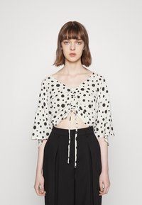 Top curto branco com bolinhas pretas, decote em V, mangas curtas tipo sino e cordão frontal para franzir. Combinado com calças pretas de cintura alta.