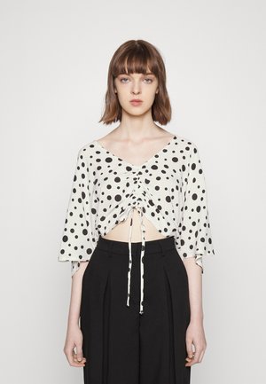 Top curto branco com bolinhas pretas, decote em V, mangas curtas tipo sino e cordão frontal para franzir. Combinado com calças pretas de cintura alta.