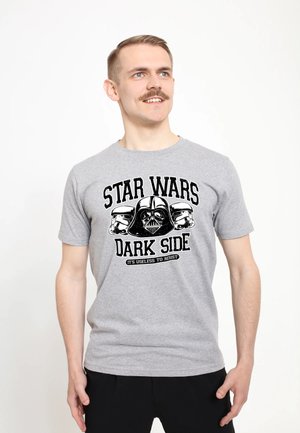 Man met snor die een grijs Star Wars Dark Side T-shirt draagt met afbeeldingen van Darth Vader en stormtrooperhelmen, glimlacht en kijkt naar rechts.