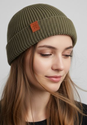 Green ribbed knit beanie with a brown leather logo patch. Designed for warmth, it features a snug fit and textured surface.
