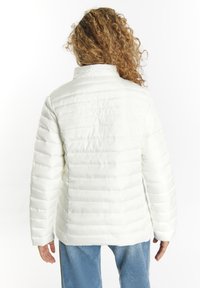 Hvid pufferjakke med vandrette sømme, højt krave og lange ærmer, lavet af skinnende materiale. Bæres over blå jeans.