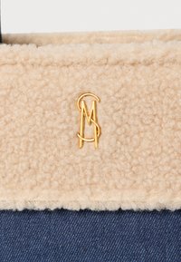 Fleece bej cu textură în partea de sus, având un logo auriu, contrastând cu o bază din denim bleumarin. Dispune de un material moale și confortabil și un design simplu.