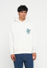 Hvit hettegenser med frontlomme, med en teal grafisk logo på brystet. Stoffet ser mykt ut med snøring i hetten. Kombinert med blå jeans.