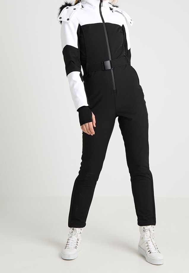 Femme portant une combinaison de ski ajustée noire et blanche avec capuche et ceinture, associée à des baskets montantes blanches, debout devant un fond uni.