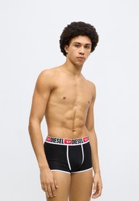 Boxeri Diesel negri cu accente albe și o talie cu brand. Modelul are o constituție medie și păr creț, fiind prezentat în picioare pe un fundal simplu.