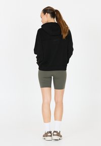 Zwarte hoodie met een relaxte pasvorm en een capuchon, gecombineerd met olijfgroene fietsshorts. Schoenen zijn bruine sneakers, waarmee een casual sportieve uitstraling wordt gecreëerd.