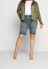 Nainen, jolla on päällään oliivinvihreä mokkatakki, valkoinen vihrein kirjailuin koristeltu paita, happopesussa käsitellyt denimshortsit ja beiget korkokengät.