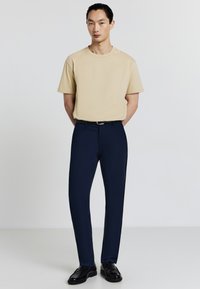 Uomo in piedi con le mani dietro la schiena, indossa una camicia beige a maniche corte, pantaloni blu navy con cintura e scarpe eleganti nere, contro uno sfondo semplice.