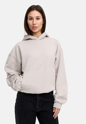 Light beige oversized hoodie with a front pocket, ribbed cuffs, and a drawstring-less hood, paired with dark denim jeans.