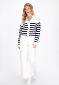 Gestreifter Cardigan in Creme und Marineblau, mit V-Ausschnitt, Knopfverschluss und weicher Textur. Kombiniert mit weißen weiten Hosen und weißen Schuhen.