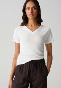 Weißes V-Ausschnitt-T-Shirt mit kurzen Ärmeln, gerippter Textur und tailliertem Design, kombiniert mit locker sitzenden dunklen Hosen mit elastischem Bund.