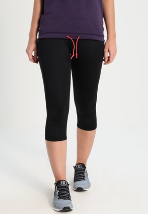 Personne portant des leggings noirs courts avec un cordon rouge et des chaussures de sport grises, debout sur un fond clair.