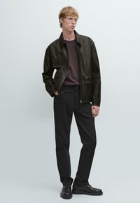 Bruine leren jas met een kraag, twee voorzakken, gecombineerd met donkere broeken en zwarte schoenen. Model staat tegen een lichte achtergrond.