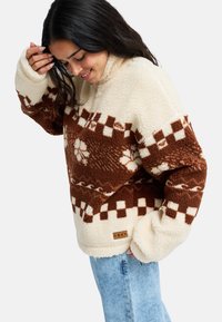 Pullover in pile marrone e crema con motivi floreali e a quadretti. Presenta un collo alto e un'etichetta con logo Roxy. Tessuto morbido e testurizzato.
