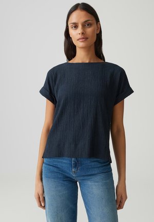 Femme aux longs cheveux foncés portant un haut court noir texturé et un jean bleu, debout devant un fond clair uni.