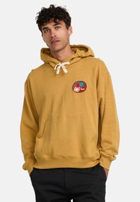 Mustard yellow hoodie made of soft cotton, featuring a front pocket and a red/green circular graphic patch with spiral designs.