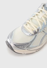 Sportowy sneaker z siateczkową cholewką w kolorze kremowym, srebrnymi akcentami i jasnoniebieskimi detalami. Posiada okrągłe sznurowadła oraz teksturowaną szarą podeszwę zewnętrzną.