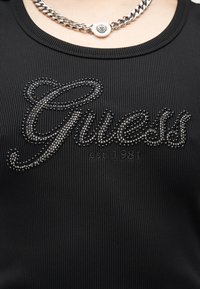 Haut en tissu côtelé noir avec l'inscription "Guess est. 1981" en noir et clous métalliques, porté avec un collier chaîne argentée avec un pendentif rond.