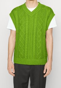 Gilet vert en tricot avec un motif torsadé et un col en V. Présente des manches courtes et un ourlet côtelé, porté sur un T-shirt blanc.