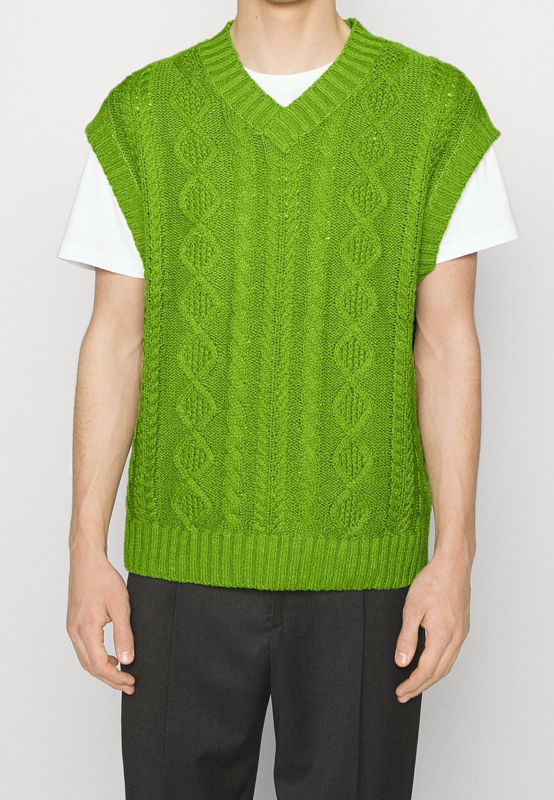 Gilet vert en tricot avec un motif torsadé et un col en V. Présente des manches courtes et un ourlet côtelé, porté sur un T-shirt blanc.