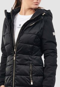 Giacca a vento nera con cappuccio, texture trapuntata, chiusura frontale con zip e due tasche con zip. Presenta dettagli in metallo.
