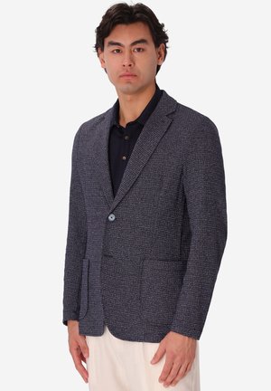 Uomo che indossa un blazer grigio scuro strutturato sopra una camicia nera con bottoni e pantaloni beige chiaro, in piedi davanti a uno sfondo bianco semplice.