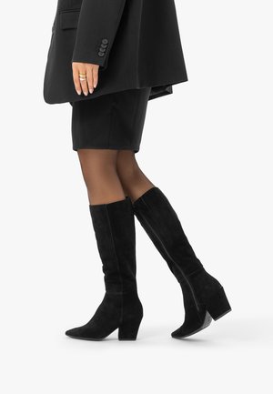 Femme portant des collants marron transparents, des bottes en daim noires montantes à talons carrés, une jupe noire et un blazer, marchant sur un fond blanc.