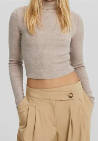 Nadmuchany, jasnobrązowy top w kroju crop z długimi rękawami i wysokim dekoltem, zestawiony z beżowymi spodniami z plisami z detalem w postaci guzika.