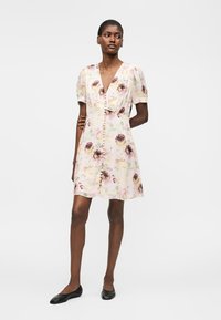 Ženska nosi kratko, svetlo cvetlično obleko z nabranimi rokavi in gumbi spredaj, v kombinaciji s črnimi ravnimi čevlji, stoji pred enobarvnim ozadjem.