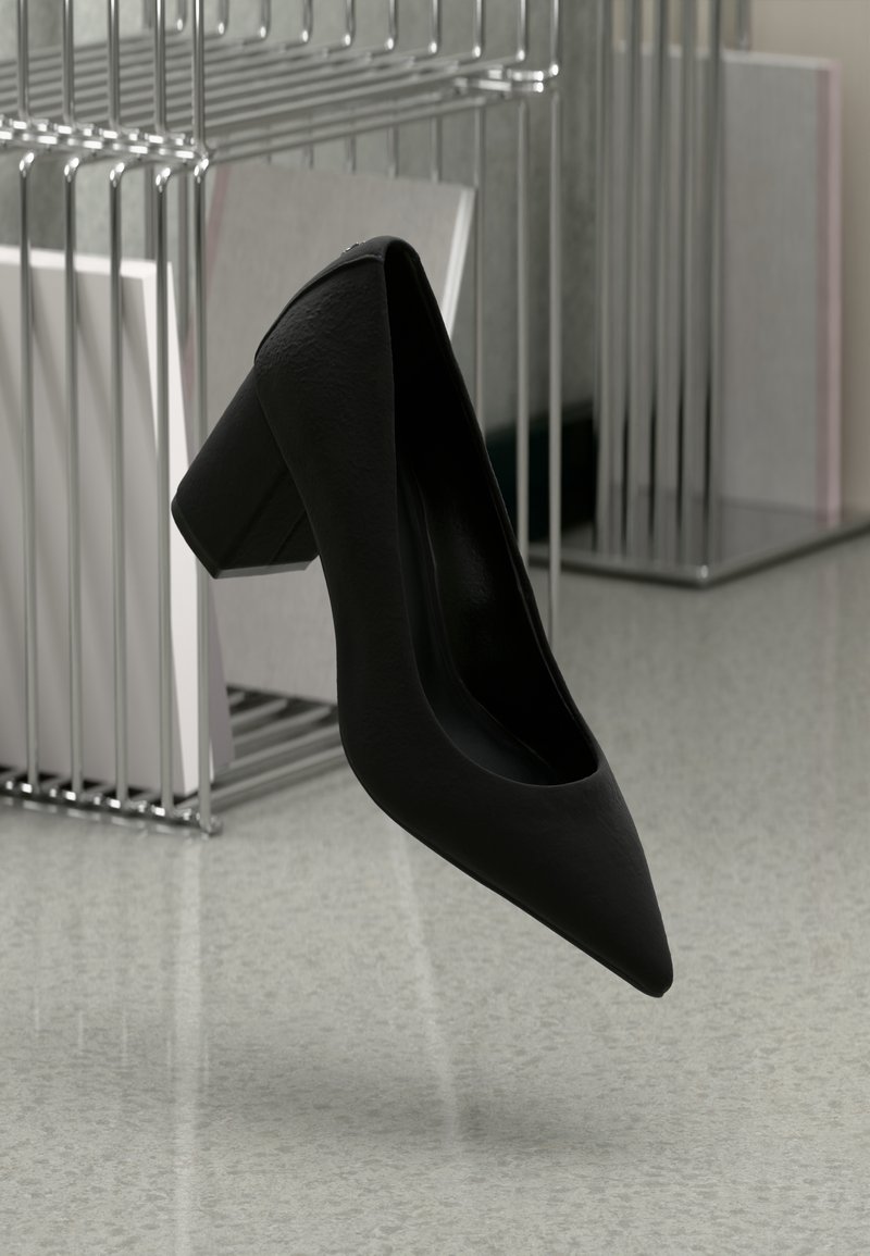 Escarpin noir à bout pointu avec talon bloc suspendu au-dessus d'un sol moucheté, porte-revues en métal avec des livres en arrière-plan.