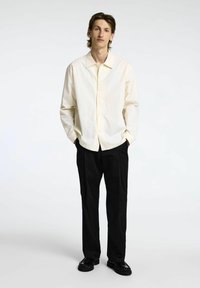 Camicia button-up di colore crema chiaro con colletto classico, abbinata a pantaloni neri e scarpe nere. L'outfit presenta una vestibilità rilassata e un design minimal.