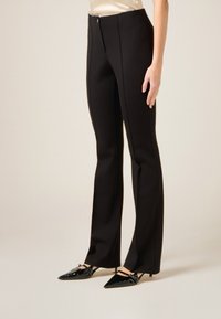 Pantalones negros a medida con un ajuste depurado, confeccionados en una tela lisa, con cremallera frontal y detalles de costuras verticales. Combinados con tacones negros puntiagudos.