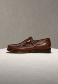 Mocassin en cuir marron avec une bride à boucle, des coutures façon mocassin et un petit talon sur une surface claire avec un fond uni.