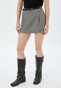 Gonna mini in bianco e nero a motivo houndstooth con pieghe, abbinata a stivali in pelle nera al ginocchio con fibbie e finitura testurizzata.