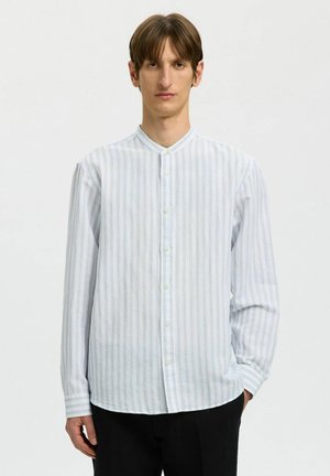 Jeune homme aux cheveux bruns portant une chemise blanche boutonnée avec de fines rayures verticales claires et un pantalon noir sur fond clair uni.