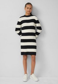 Schwarz-weiß gestreiftes Strickkleid mit langen Ärmeln und Rundhalsausschnitt, kombiniert mit weißen Sneakern, vor einem schlichten Hintergrund stehend.