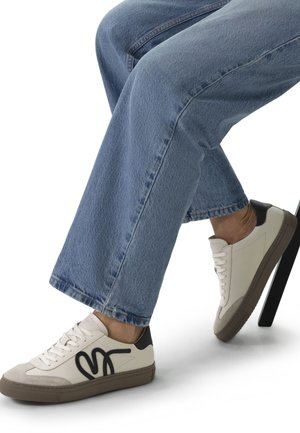 Persoon die lichtblauwe spijkerbroek en witte sneakers met zwarte en beige accenten draagt, zittend op een stoel tegen een witte achtergrond.