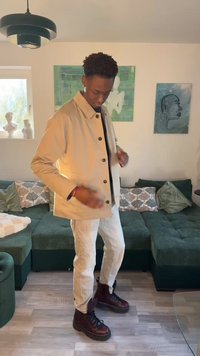 Νεαρός άνδρας που ρυθμίζει ένα μπεζ σακάκι, όρθιος στο σαλόνι με πράσινο καναπέ, μαξιλάρια με σχέδια, τοιχογραφίες και ξύλινο πάτωμα.